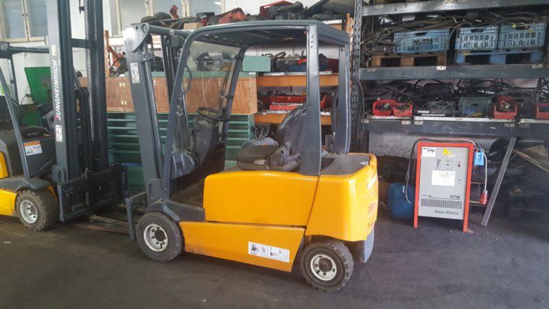 Żółty wózek widłowy elektryczny Jungheinrich na tle magazynu z regałami i ładowarką akumulatorów.