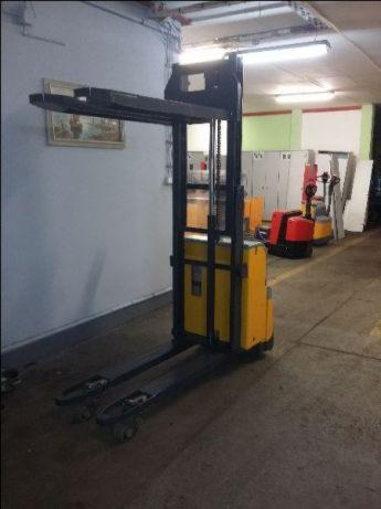 Żółty, elektryczny wózek paletowy stojący w magazynie, z widocznymi innymi wózkami w tle.