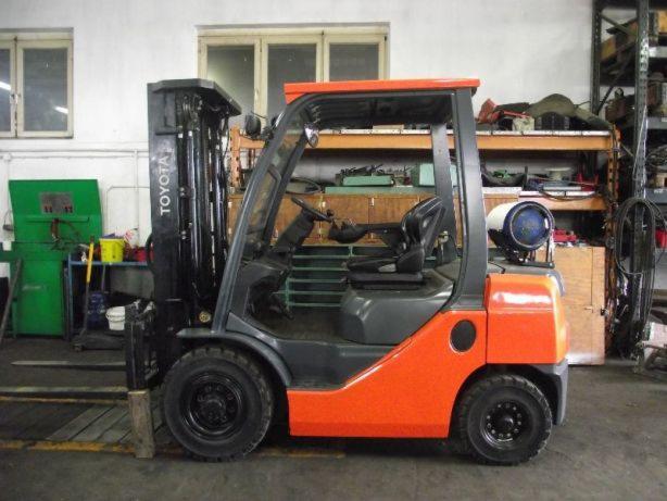 Pomarańczowo-szary wózek widłowy Toyota z butlą gazową z tyłu, stojący w warsztacie na tle regałów i narzędzi.