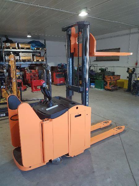 Pomarańczowy wózek widłowy elektryczny stojący w magazynie, widoczne półki z różnymi przedmiotami w tle.