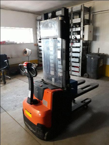 Pomarańczowo-czarny elektryczny wózek paletowy z przezroczystą osłoną na widłach, stojący w magazynie z metalowymi regałami w tle.