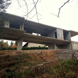 Surowy, betonowy szkielet dwukondygnacyjnego domu w trakcie budowy, widoczny z dołu na tle pochmurnego nieba i drzew.