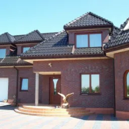 Elewacja domu z cegły klinkierowej z brązowymi oknami i drzwiami. Widoczne schody wejściowe, rzeźba orła i brama garażowa.