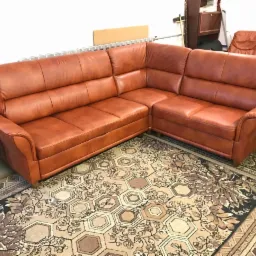 Narożnik obity jasnobrązową skórą, ustawiony na wzorzystym dywanie w pomieszczeniu z meblami w tle.