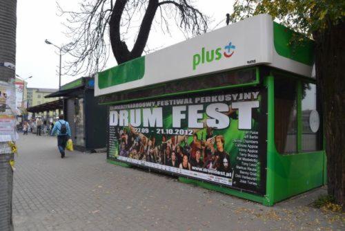 Kiosk z plakatem Drum Fest 2012, logo Plus na górze, zielone akcenty, chodnik i przechodzień w tle. Plakat z listą artystów i datą wydarzenia.