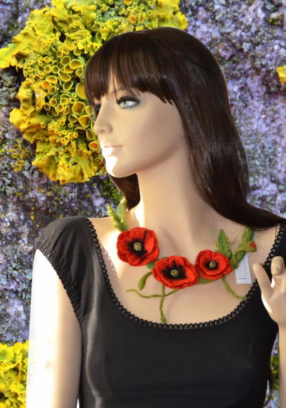 Manekin w czarnej sukience prezentuje naszyjnik z filcowanymi makami na tle abstrakcyjnej ściany w odcieniach żółci i fioletu. Nietypowe tło dodaje zdjęciu unikalności.