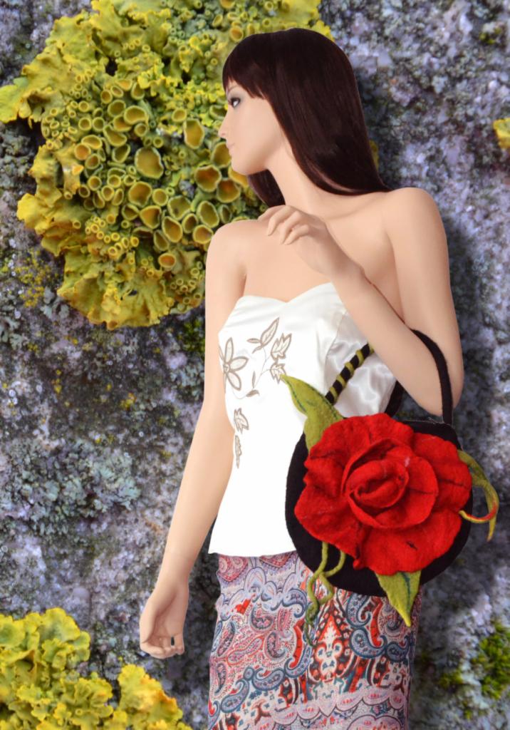 Manekin w białym gorsecie z haftem, trzymający czarną torebkę z czerwoną różą na tle skały porośniętej żółtymi porostami. Nietypowe tło i stylizacja.