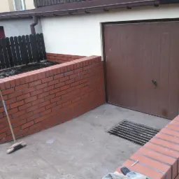 Klinkierowa obudowa rabaty kwiatowej i fragment elewacji z brązową bramą garażową. Widoczna studzienka kanalizacyjna i narzędzia glazurnicze.