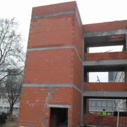 Surowa konstrukcja budynku z czerwonej cegły, widoczne betonowe stropy, w tle drzewo i budynki. Etap budowy w trakcie realizacji, widoczny pracownik w odblaskowej kamizelce.