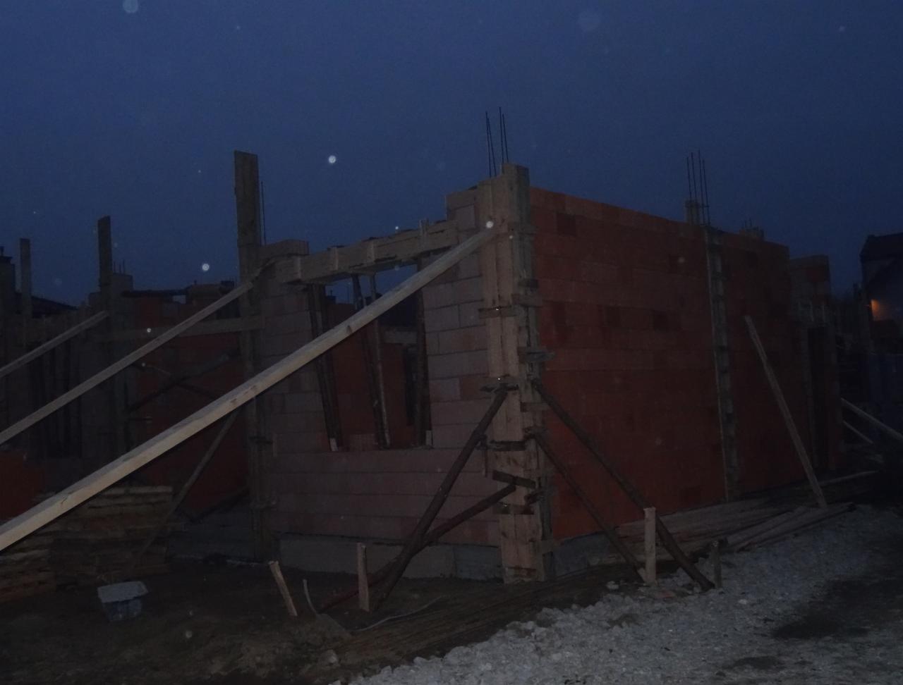 Budowa domu z czerwonej cegły w trakcie realizacji, widoczne drewniane podpory i zbrojenia, nocne ujęcie.