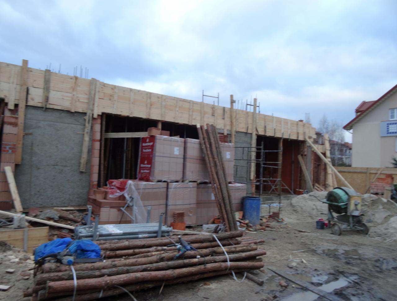 Budowa domu w trakcie wznoszenia ścian z cegły, widoczne szalunki, materiały budowlane i betoniarka. Prace murarskie w toku, pochmurne niebo w tle.