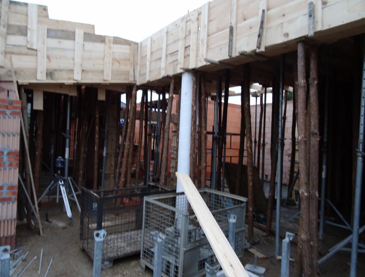 Szczegółowy widok szalunku stropu z drewnianymi podporami i metalowymi koszami na budowie, z ceglaną ścianą w tle i niwelatorem na statywie.