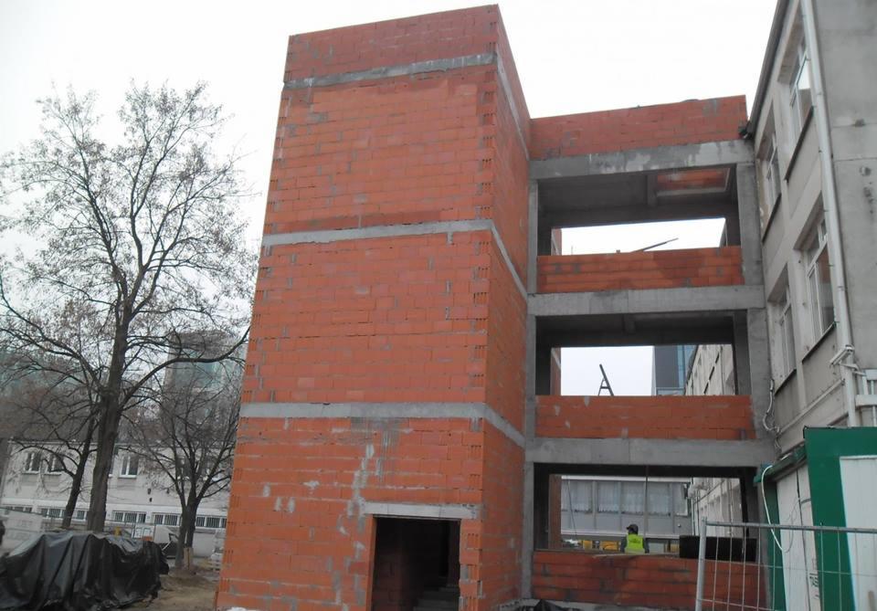 Surowa konstrukcja budynku z czerwonej cegły, widoczne betonowe stropy, w tle drzewo i budynki. Etap budowy w trakcie realizacji, widoczny pracownik w odblaskowej kamizelce.