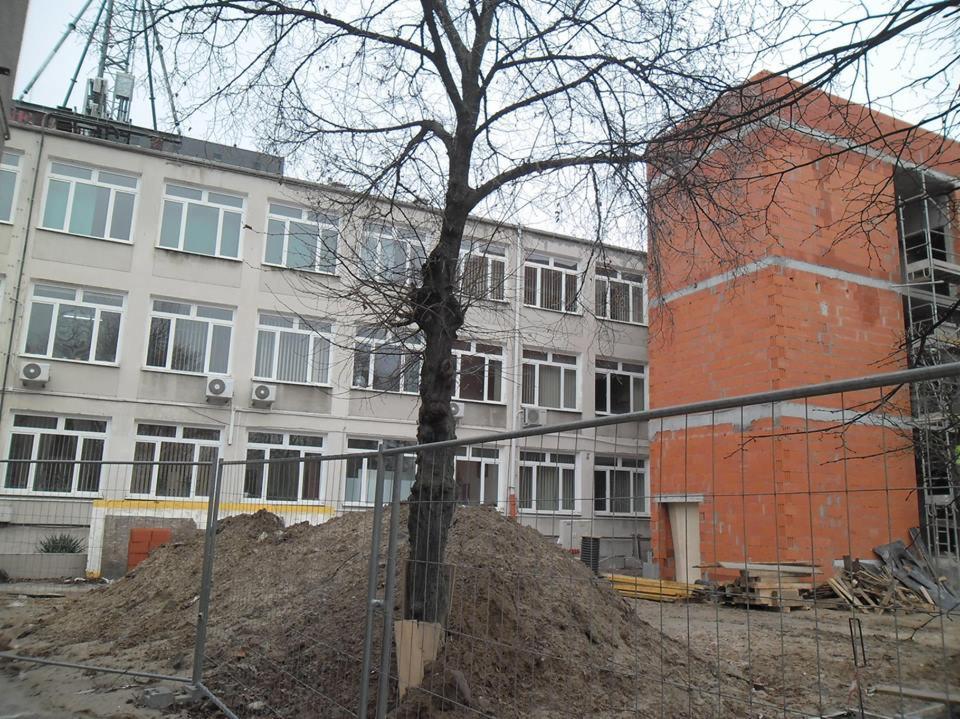 Plac budowy z ogrodzeniem tymczasowym, pryzma ziemi, budynek z klimatyzacją i surowy budynek z cegły kratówki w tle. Drzewo bez liści na pierwszym planie.