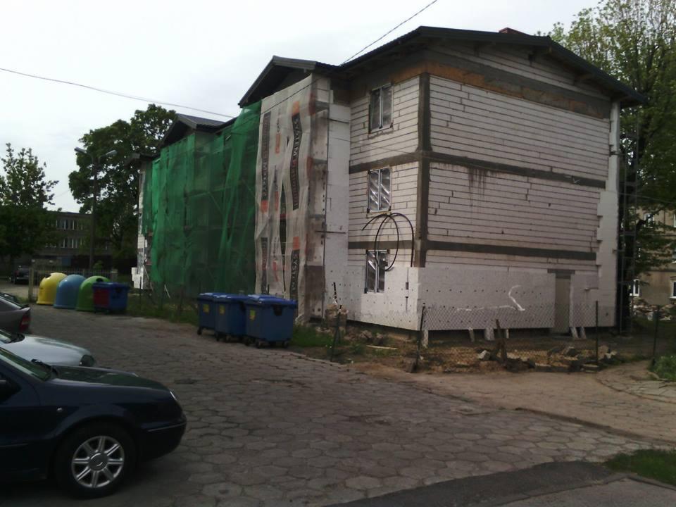 Dwupiętrowy budynek w trakcie ocieplania, z widocznym rusztowaniem i siatką ochronną. Część elewacji już ocieplona styropianem, druga w trakcie prac. Widoczne kontenery na śmieci.