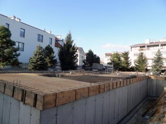 Zbrojenie fundamentów pod budynek wielorodzinny, widoczne szalunki i stalowe pręty. W tle zabudowa miejska i drzewa w słoneczny dzień.