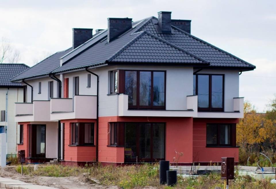 Dwukondygnacyjny dom szeregowy z ciemną dachówką i jasną elewacją, z balkonami i dużymi oknami, otoczony zielenią i widoczny z perspektywy ulicy.