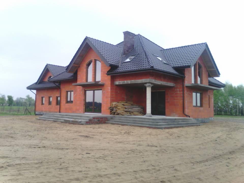 Surowy, dwukondygnacyjny dom z czerwonej cegły z ciemnym dachem, betonowymi schodami i kolumną. W tle drzewa i pole. Częściowo widoczne materiały budowlane.