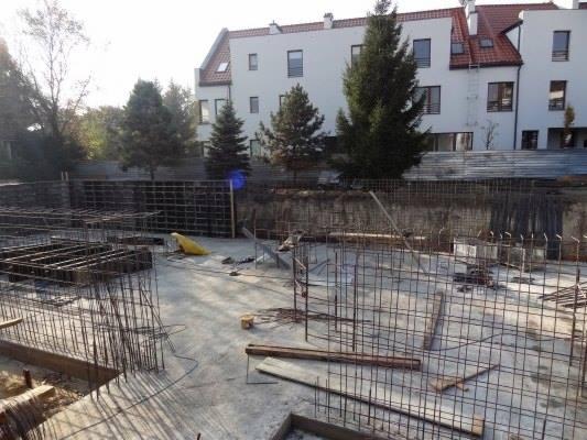 Zbrojenie fundamentów pod budynek mieszkalny. Widoczne stalowe pręty tworzące szkielet konstrukcji. W tle budynek z czerwonym dachem i drzewa.