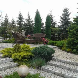 Ogród z kamienną ścieżką i schodami, otoczony zielenią i drzewami iglastymi. W tle zestaw mebli ogrodowych, tworzący zaciszny kącik wypoczynkowy.