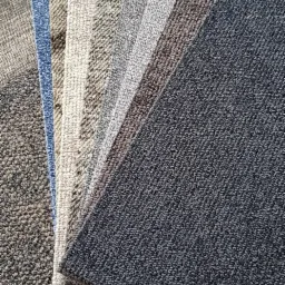 Próbki wykładzin dywanowych w różnych odcieniach szarości i błękitu, ułożone wachlarzowo na tle wykładziny o drobnej strukturze.