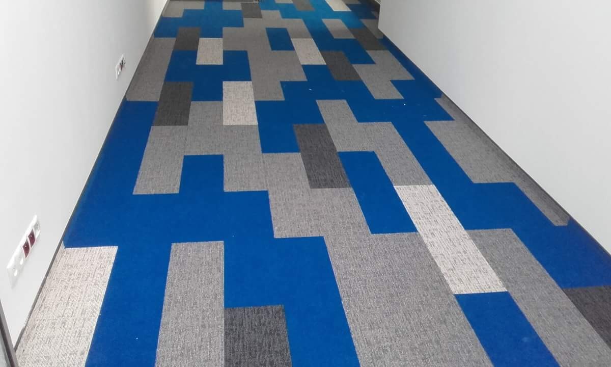 Wzorzysta podłoga wyłożona wykładziną dywanową w odcieniach niebieskiego, szarego i białego, ułożona w geometryczny wzór wzdłuż korytarza z białymi ścianami.