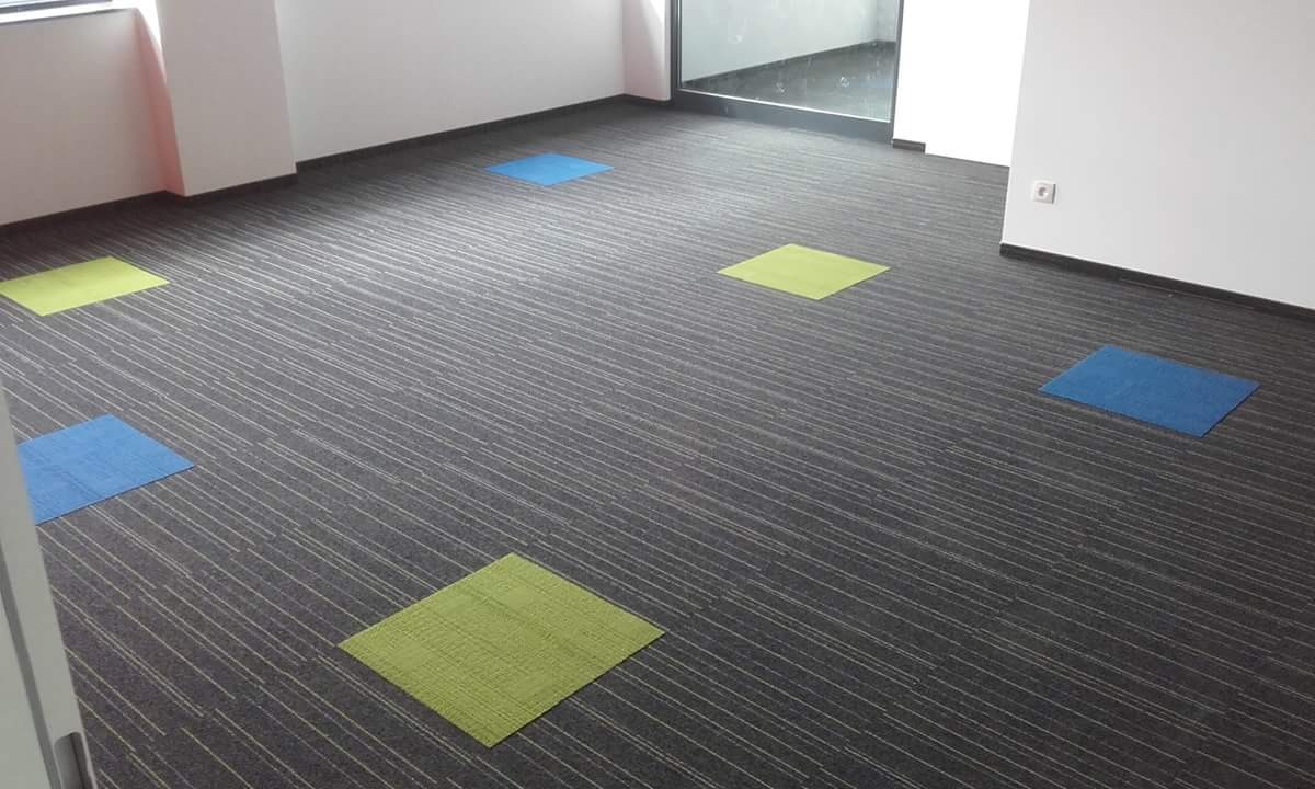 Biuro z szarą wykładziną w paski i kolorowymi kwadratami dywanowymi w odcieniach niebieskiego i zielonego.