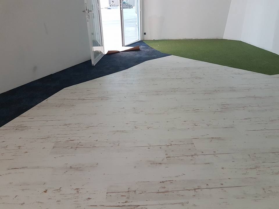 Wnętrze z trzema różnymi rodzajami pokrycia podłogowego: panele imitujące bielone drewno, ciemna wykładzina dywanowa i sztuczna trawa, widok na otwarte drzwi balkonowe.