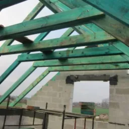 Konstrukcja dachu z impregnowanej tarcicy w kolorze zielonym, wznoszona na ścianach z betonu komórkowego. Widok na otwarty krajobraz przez okno w murze.