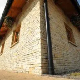 Elewacja budynku wykończona jasnym, nieregularnym kamieniem, z widocznymi oknami i drewnianymi elementami pod dachem. Ujęcie z dołu, podkreślające strukturę kamienia.