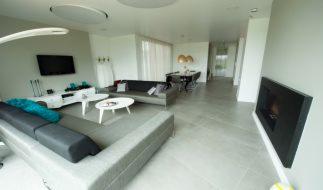 Nowoczesne wnętrze salonu z szarą sofą narożną, białymi stolikami kawowymi i kominkiem. Minimalistyczny design z telewizorem na ścianie i dużym oknem.