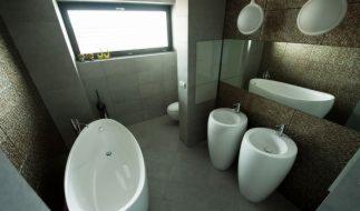 Nowoczesna łazienka z wolnostojącą wanną i umywalkami na postumentach, szara kolorystyka, okno, lustro. Minimalistyczny design, oświetlenie punktowe.