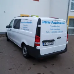 Biały samochód dostawczy firmy budowlanej Peter Pawlowski z niebieskim napisem na tle budynku z kolorowymi ramami okiennymi.