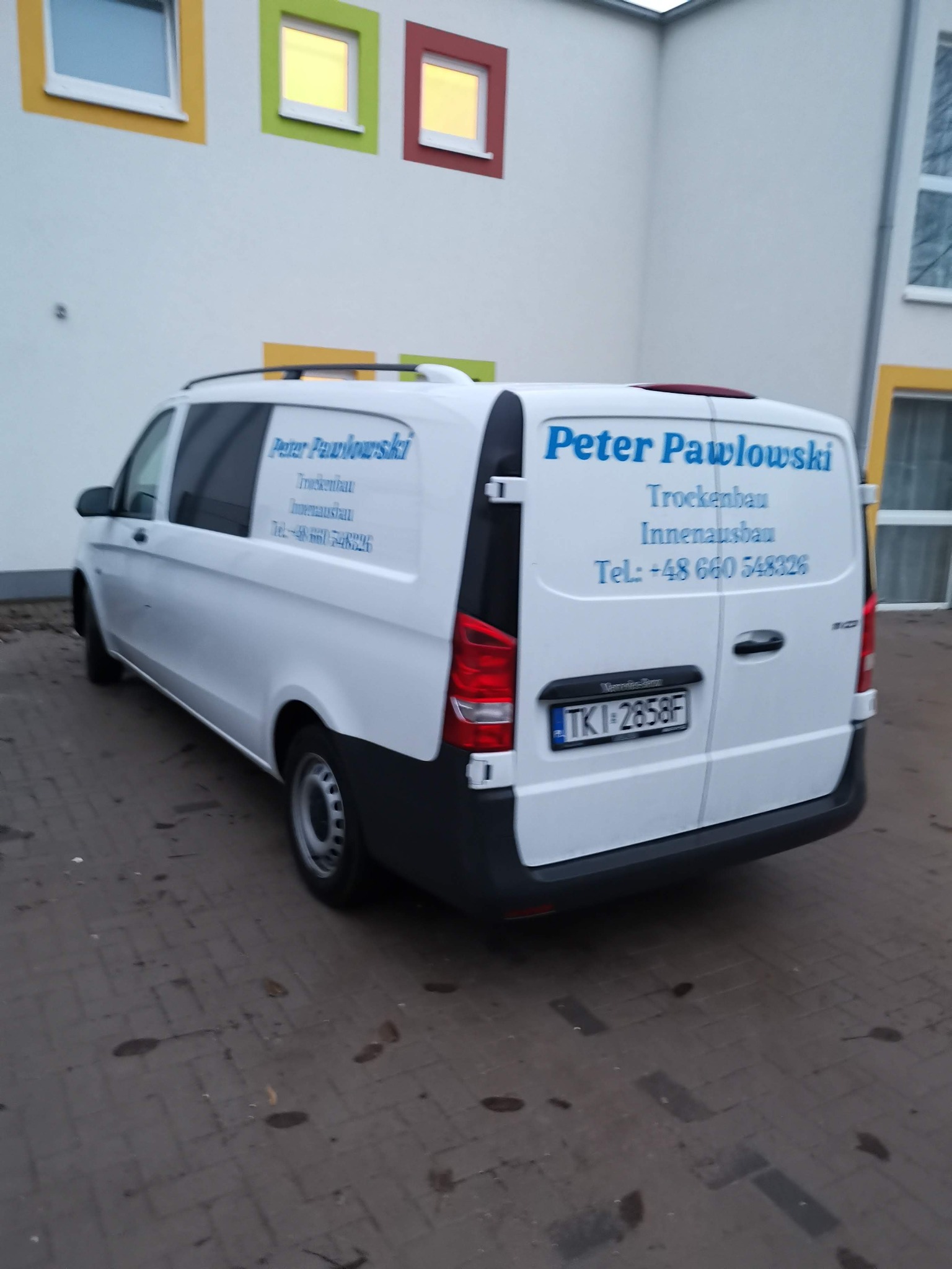 Biały samochód dostawczy firmy budowlanej Peter Pawlowski z niebieskim napisem na tle budynku z kolorowymi ramami okiennymi.