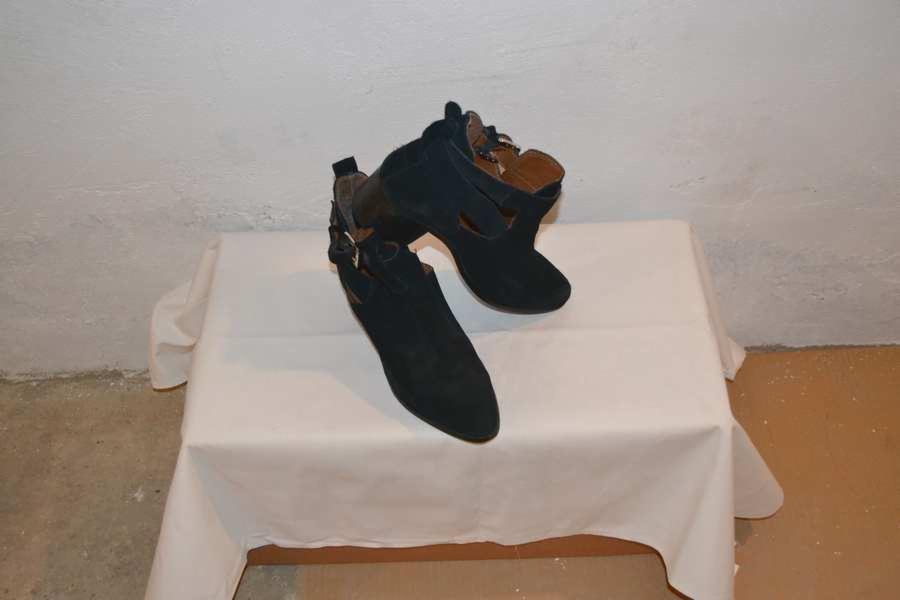 Para czarnych, zamszowych botków damskich z wycięciami, ustawiona na białym obrusie na tle surowej, białej ściany. Buty posiadają brązowe wnętrze i zapięcie na sprzączkę.