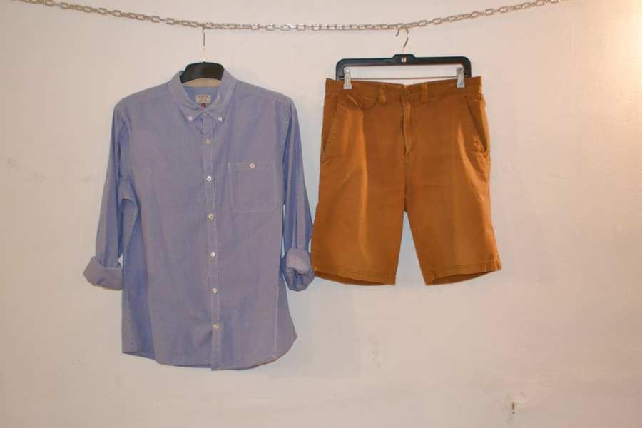 Używana, błękitna koszula z długim rękawem i brązowe szorty na wieszakach, zawieszone na łańcuchu na tle białej ściany. Odzież z drugiej ręki.