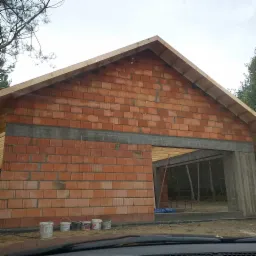 Wznoszenie ścian zewnętrznych budynku z czerwonej cegły, widoczny betonowy nadproże nad otworem drzwiowym i drewniana konstrukcja dachu w surowym stanie, kilka wiader z farbą u podstawy ściany.