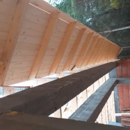 Konstrukcja drewniana więźby dachowej w trakcie budowy, widoczne krokwie, łaty i mur z czerwonej cegły, ujęcie od dołu w kierunku korony drzew