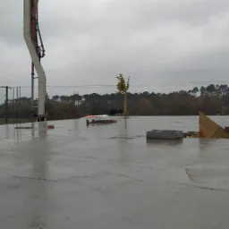 Wylany, mokry fundament domu z wystającymi prętami zbrojeniowymi, widoczna pompa do betonu, szalunek, bloczki betonowe i drzewo w tle.