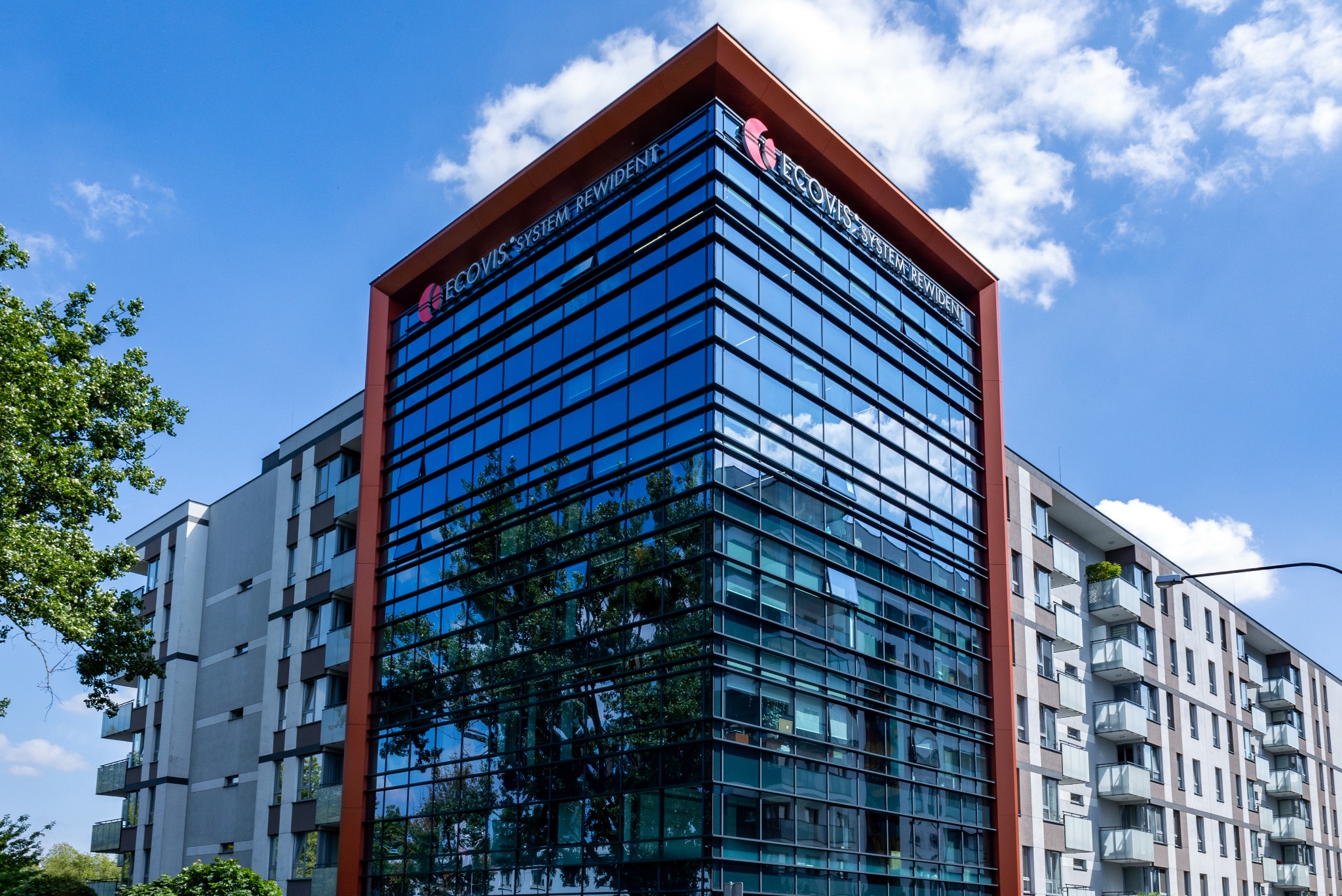 Nowoczesny biurowiec z logo Ecovis System Rewident na tle błękitnego nieba i fragmentów innych budynków. Szklana fasada odbija zieleń drzew.