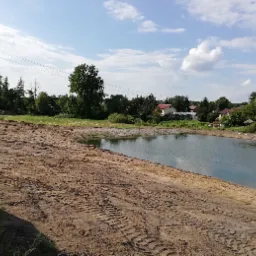 Nowo wykopany staw z brzegami z ubitej ziemi, widoczne ślady opon maszyn budowlanych, w tle zabudowania wiejskie i drzewa.