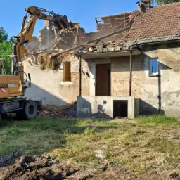 Żółta koparka marki Liebherr rozbiera stary, murowany dom z czerwonym dachem w słoneczny dzień, z widocznymi resztkami dachu i gruzem.