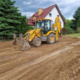 Żółta koparko-ładowarka JCB 3CX na placu budowy, wyrównująca teren przed domem z czerwoną dachówką, widoczne ślady opon na ziemi.
