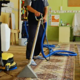 Czyszczenie dywanu w klasie szkolnej za pomocą profesjonalnego odkurzacza piorącego z żółtą obudową i niebieskimi wężami. Widoczne biurko i krzesło w tle.