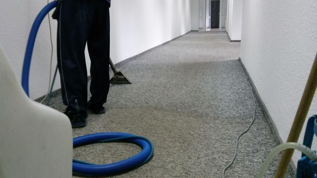 Czyszczenie tekstylnej podłogi w korytarzu biurowym za pomocą odkurzacza piorącego. Widoczny fragment urządzenia i węże. Szara wykładzina w jasnym wnętrzu.
