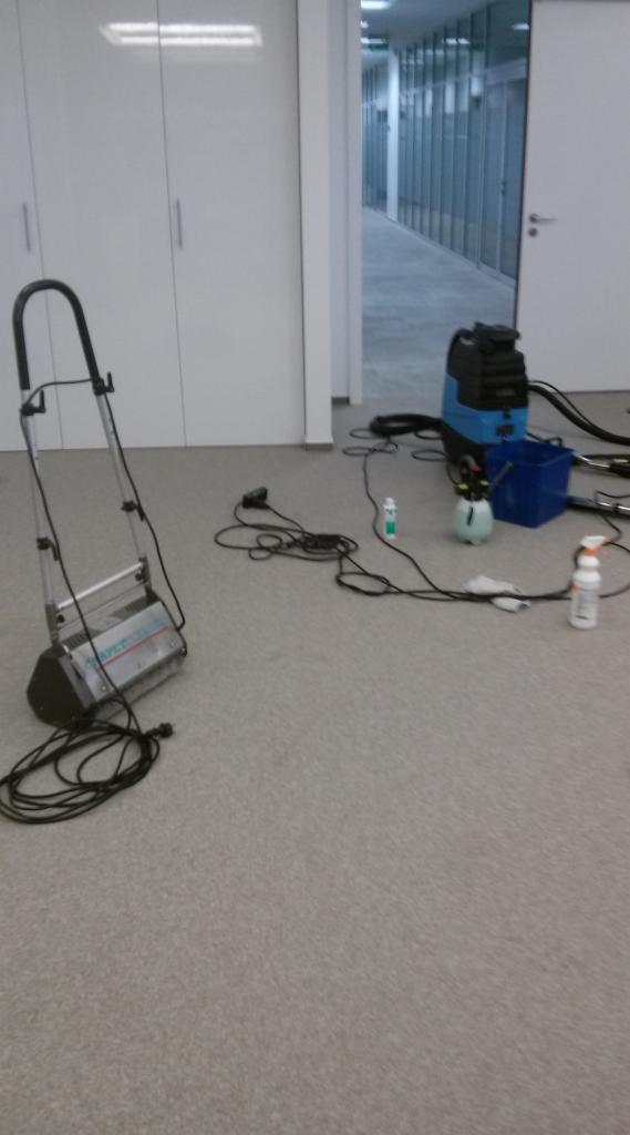 Maszyna do czyszczenia dywanów i akcesoria na jasnej wykładzinie w biurze. W tle białe szafy i korytarz z przeszklonymi ścianami.