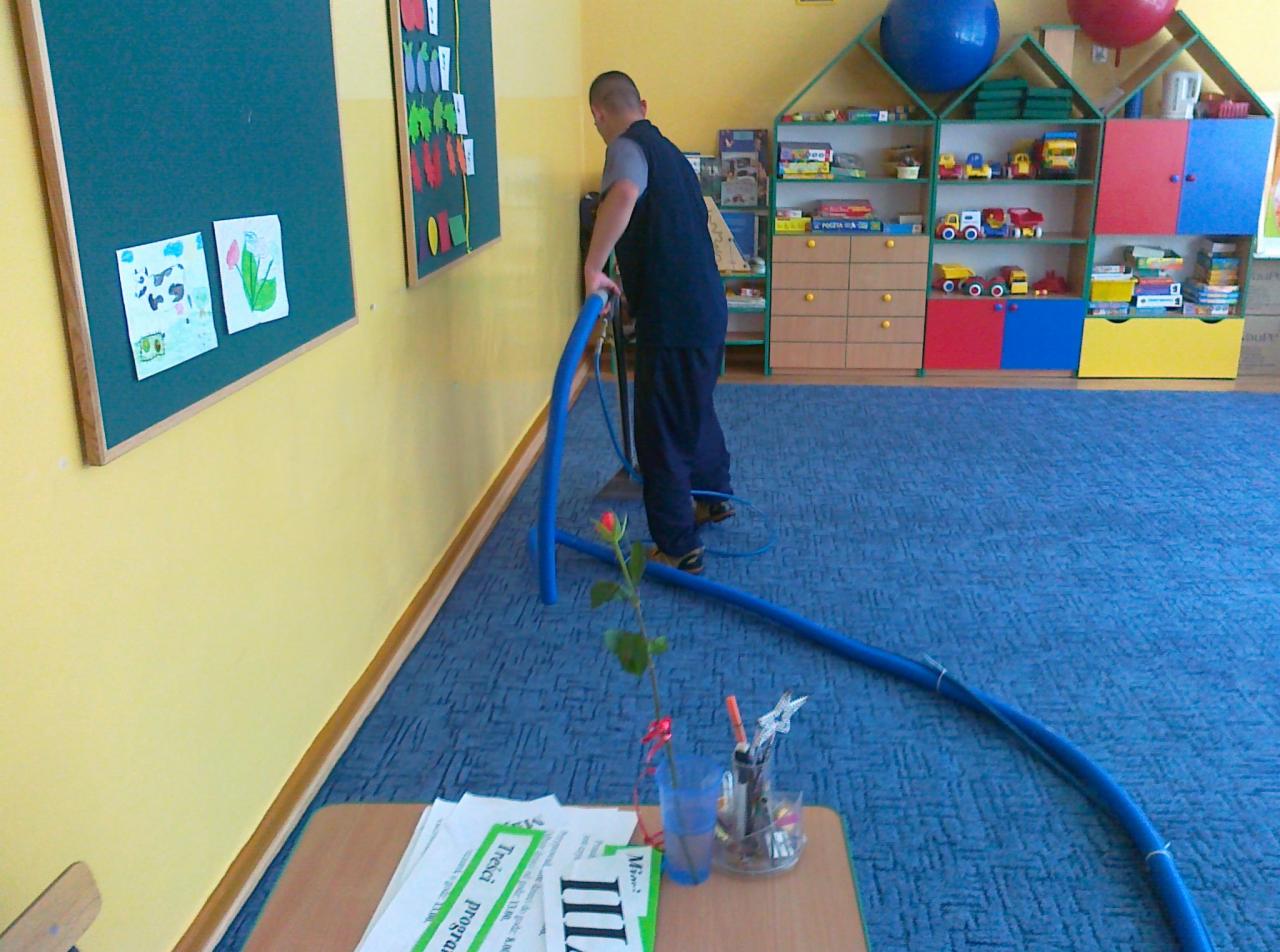 Mężczyzna czyści niebieską wykładzinę w pokoju dziecięcym odkurzaczem piorącym. Na pierwszym planie stolik z kwiatem i programem zajęć.