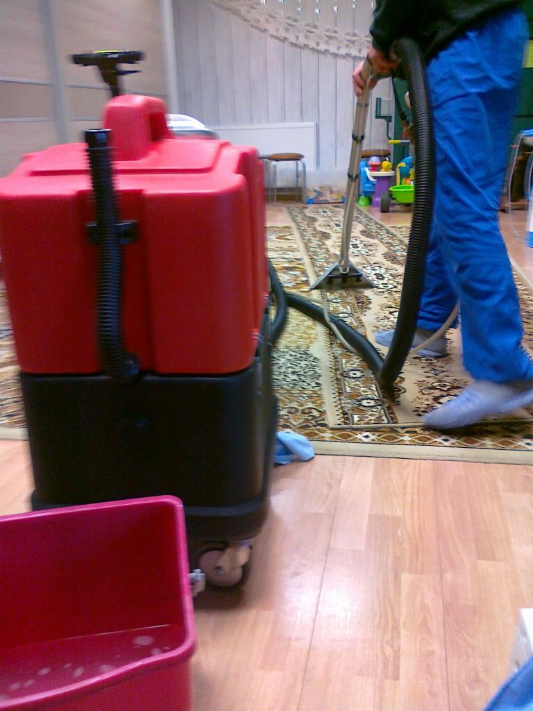 Czyszczenie dywanu za pomocą profesjonalnego odkurzacza piorącego. Widoczny fragment dywanu, odkurzacz w kolorze czerwono-czarnym i osoba w niebieskich spodniach.