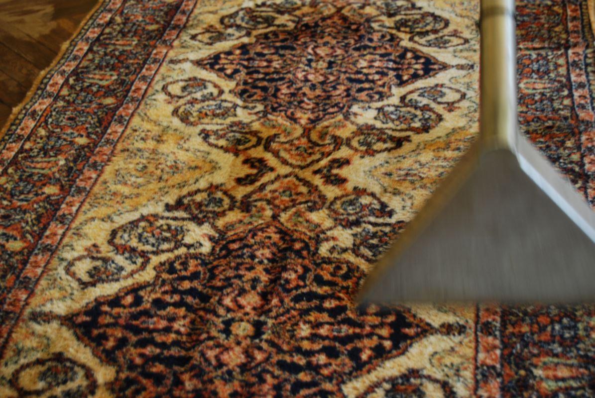 Czyszczenie wzorzystego dywanu z użyciem dyszy parowej. Widoczny fragment drewnianej podłogi. Dywan w odcieniach beżu, brązu i granatu.