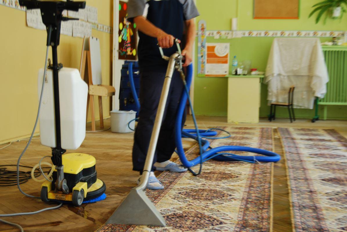 Czyszczenie dywanu w klasie szkolnej za pomocą profesjonalnego odkurzacza piorącego z żółtą obudową i niebieskimi wężami. Widoczne biurko i krzesło w tle.